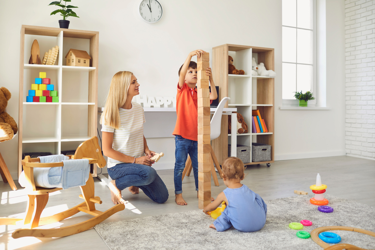 Happy Mother with Her Two Children Playing with Lots of Toys and Building Block Tower at Home